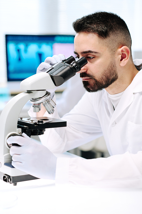Young bearded man in lab coat and gloves looking at bacteria in microscope while sitting by his workplace in laboratory