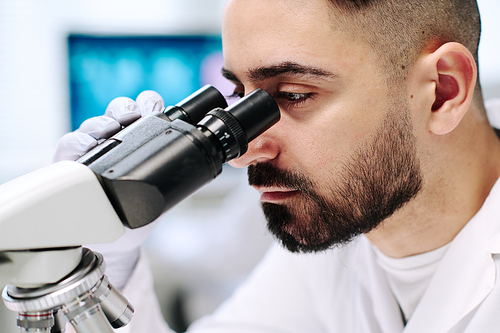 Part of face of young male researcher with microscope studying new virus while sitting by workplace against computer screen in laboratory