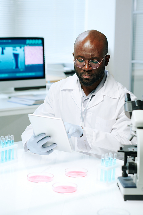African American scientist in whitecoat, gloves and eyeglasses searching for online scientific data while using tablet by workplace