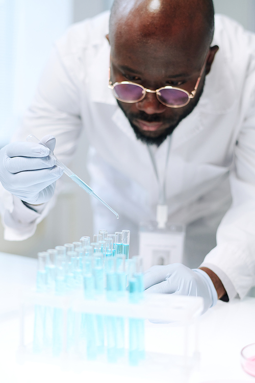 African American chemist mixing two liquid substances in chemical laboratory while dropping one of them into several flasks