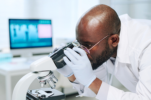 Young serious black man in whitecoat and gloves working with microscope while making scientific research in laboratory