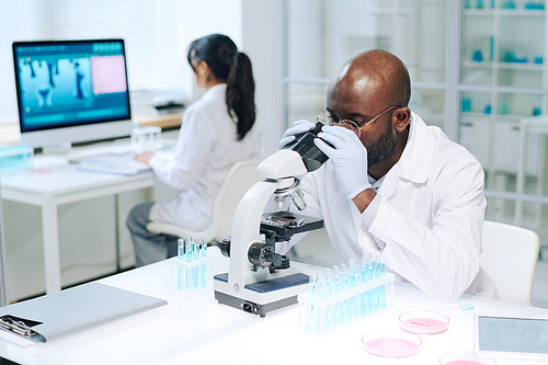 Young African American male researcher in whitecoat and gloves making scientific experiment while using microscope against female coworker