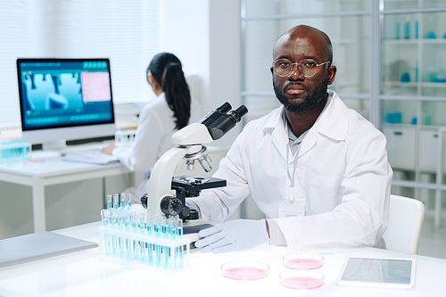 Young serious scientist in lab coat sitting by workplace in front of microscope and looking at camera during scientific work