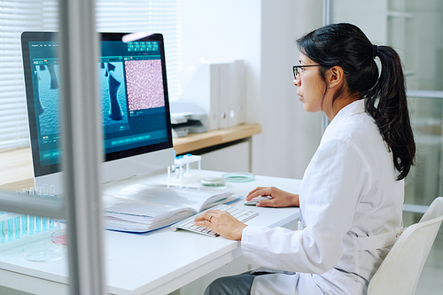 Side view of young Hispanic female researcher sitting in front of computer monitor and studying new virus in modern scientific laboratory