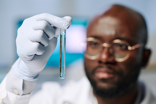 Gloved hand of African American male researcher holding flask with light blue liquid and looking at it during scientific investigation