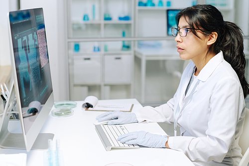 Young serious female researcher or virologist in whitecoat and gloves sitting in front of computer screen while analyzing new strain