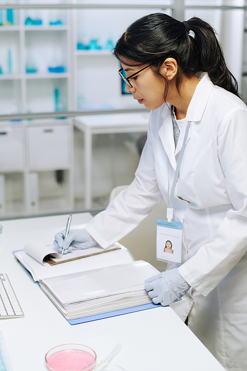 Young female biochemist in whitecoat and gloves making scientific notes in notepad while standing by desk in clinical laboratory