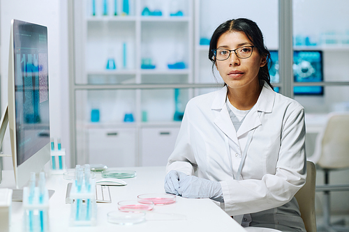 Young serious female virologist or microbiologist in whitecoat looking at camera while sittiing by workplace with test tubes and petri dishes
