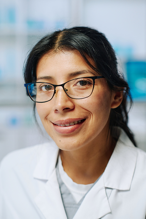 Young successful Hispanic female biochemist in eyeglasses and whitecoat looking at camera with smile while working in laboratory