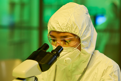 Contemporary female virologist in protective coveralls using microscope during scientific research or experiment in laboratory