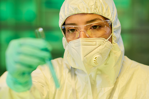 Female scientist in protective eyeglasses, respirator and biohazard suit looking at chemical sample in test tube in her gloved hand