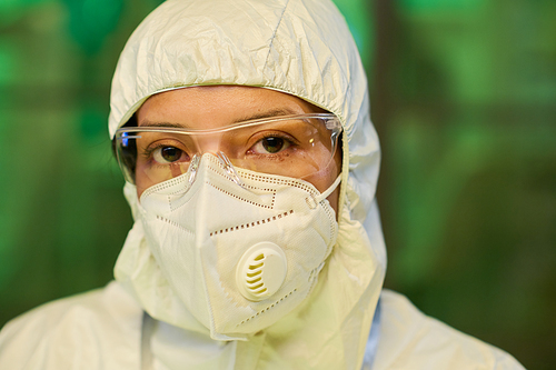 Face of young female scientist in protective eyeglasses, respirator and coveralls standing in front of camera in clinical laboratory