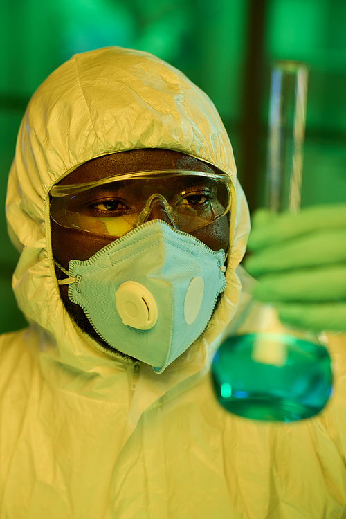 Young black man in protective coveralls looking at big test tube with blue liquid while studying characteristics of new chemical substance