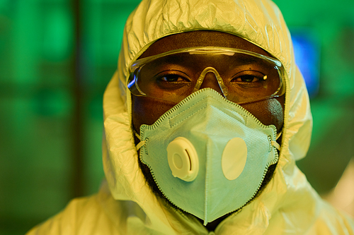 Face of African American male virologist in protective coveralls, eyeglasses and respirator standing in front of camera in laboratory