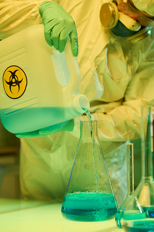 Close-up of modern scientist in protective workwear mixing liquid substances while pouring one of them into big test tube