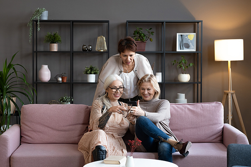 Group of mature women watching something on mobile phone and smiling sitting on sofa in the room