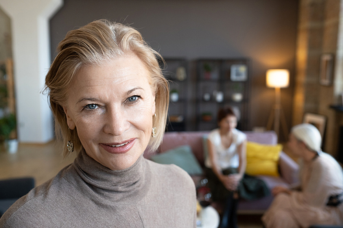 Portrait of mature woman smiling at camera with friends sitting on sofa in the background, she meeting with them at home