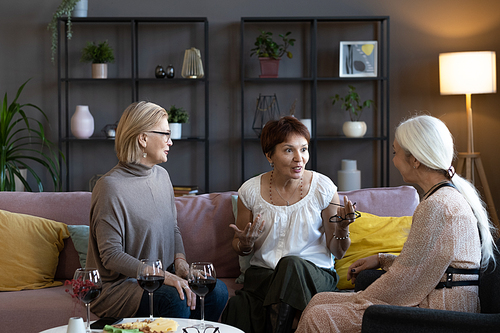 Group of mature friends sitting on sofa and talking to each other with glasses of red wine on the table in the living room