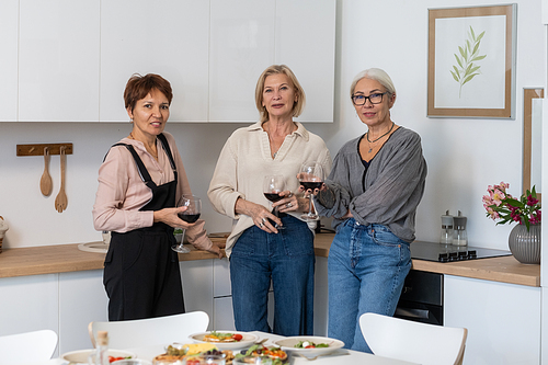 Portrait of mature friends smiling at camera while drinking red wine standing in the kitchen at home