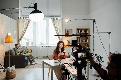 Happy young female blogger sitting by table in loft apartment during video record of commercial being shot by cameraman