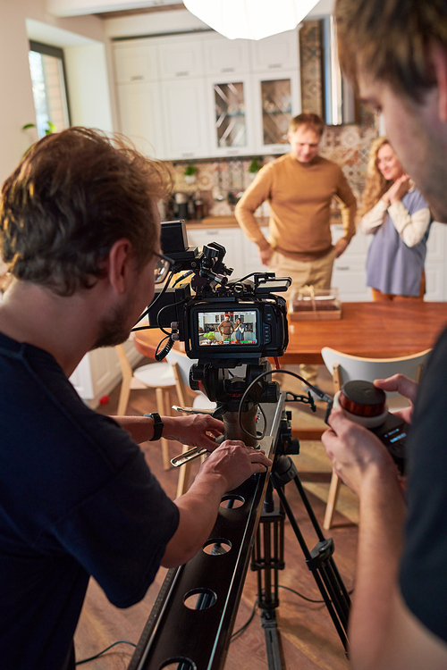 Two young videographers moving video camera along slider during shooting of commercial or cooking masterclass in the kitchen