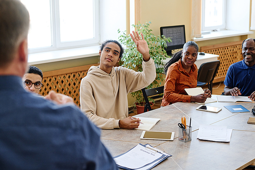 Young immigrant Black student attending language classes sitting at table raising hanf to ask teacher question