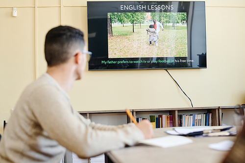 Selective focus on background shot of unrecognizable young man sitting at table watching video or presentation during English lesson