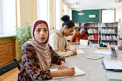 Young woman wearing hijab sitting at table with her classmates looking at camera during English language lesson