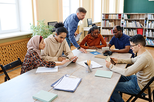 Mature Caucasian English language teacher helping immigrant students with doing task during lesson in library