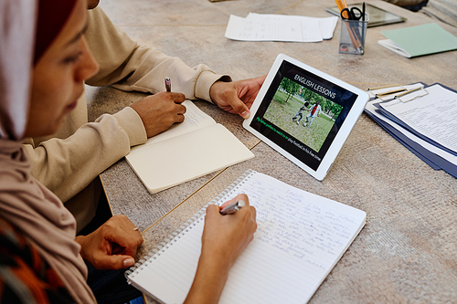 Muslim woman and her unrecognizable classmate watching education video on digital tablet and making notes during English lesson