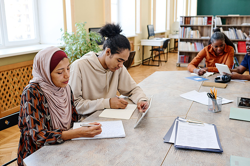 Group of multi-ethnic immigrant students sitting at table watching educational videos on digital tablet and doing task in pairs