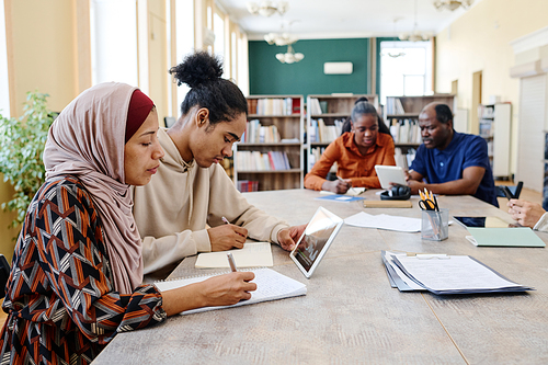 Group of ethnically diverse students sitting at table watching educational videos on digital tablet and doing writing task