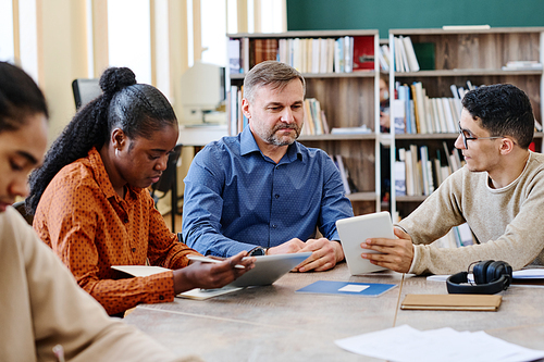 Mature language teacher sitting at table with his ethnically diverse students watching them doing exercise and answering questions