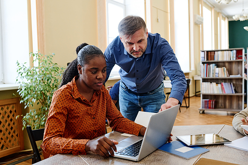 Mature Caucasian man working as teacher helping Black student to do task correctly using laptop and internet