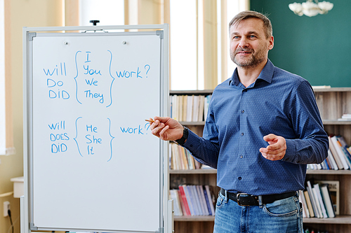 Portrait of mature Caucasian teacher wearing blue shirt and jeans standing next to whiteboard explaining English question grammar rules