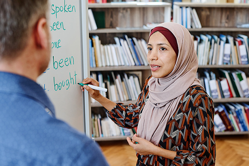 Modern Muslim woman wearing hijab standing at whiteboard doing task with irregular verbs and answering teachers question during English lesson