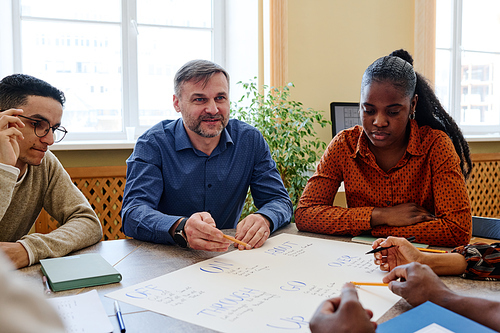 Professional English language teacher sitting among his student explaining meaning of phrasal verb written on poster