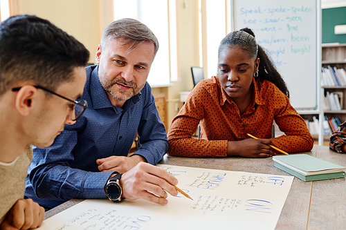 Mature English language teacher sitting at table with his students asking them questions during lesson