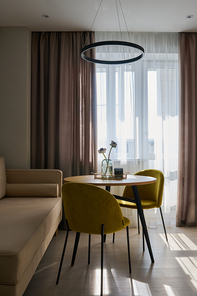 Two mustard yellow chairs standing by round table next to beige sofa against white and grey curtains on window in spacious kitchen