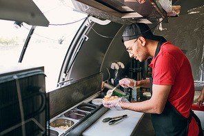 Young man in uniform and gloves cooking tasty hotdogs and other snacks in food truck