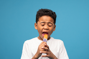 Adorable little boy of African ethnicity licking tasty icecream with taste of juicy orange while looking at you against blue background