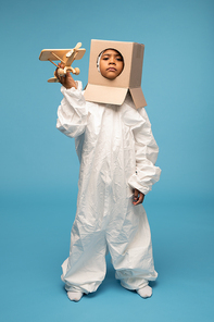 Cute funny cosmonaut in white clothes and cardboard box on his head keeping right hand by temple while playing in living-room