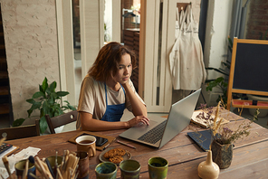 Young creative woman concentrating on network while sitting by table with handmade earthenware in large workshop