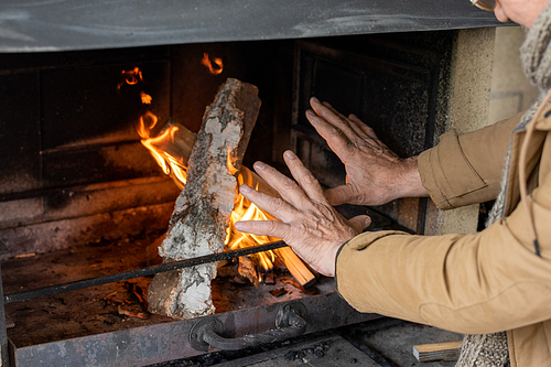 Hands of senior man in brown jacket standing by fireplace with burning firewoods while getting warm on cool autumn day in country house