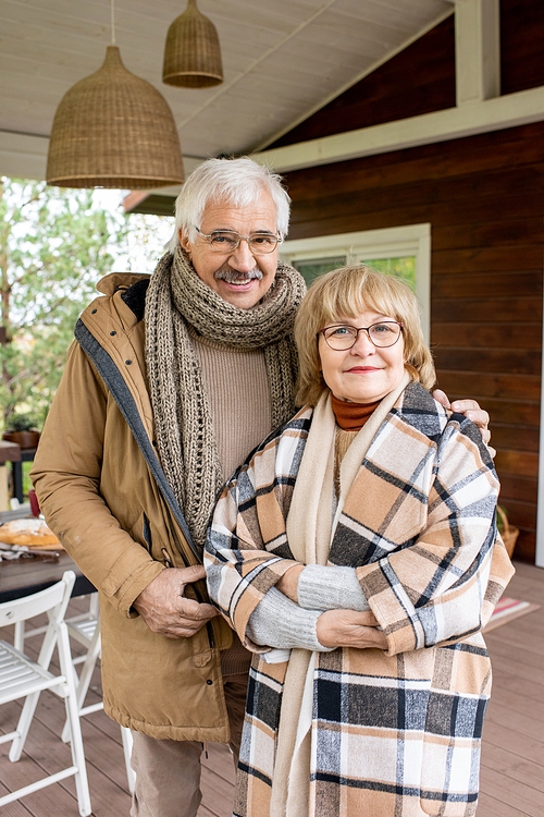 Affectionate senior husband and wife in warm casualwear looking at you with smiles while standing by their house against served table