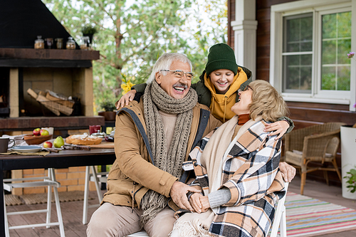 Happy cute schoolboy and his affectionate grandparents in warm casualwear laughing while sitting against house and table