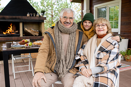 Happy cute schoolboy and his affectionate grandparents in warm casualwear looking at you with smiles against house and table