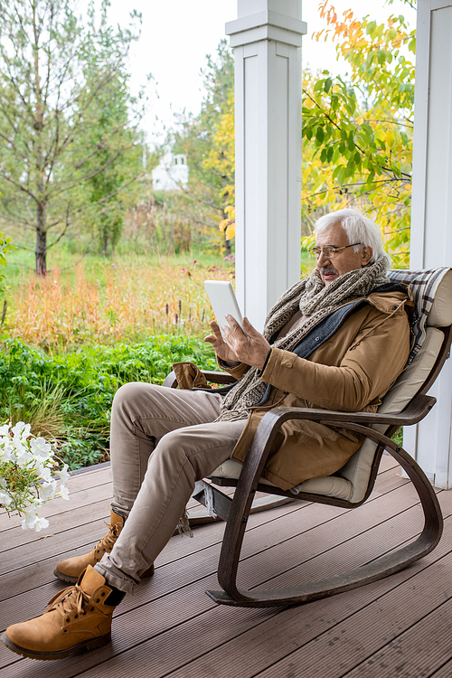 Happy senior man with white hair using tablet while sitting in rocking chair and communicating through video chat or surfing in the net