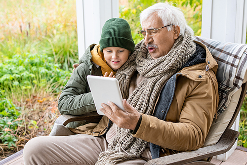 Cute schoolboy in beanie and warm jacket and his grandfather with digital tablet looking at touchscreen while discussing news outdoors