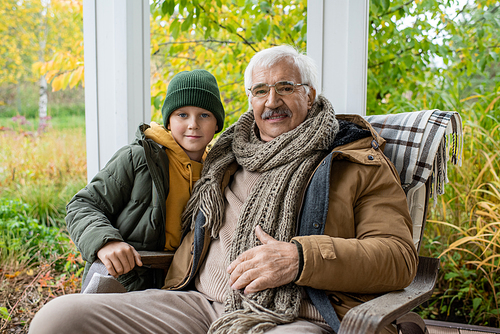 Cute schoolboy in beanie and warm jacket and his grandfather in rocking chair looking at you while relaxing against natural environment